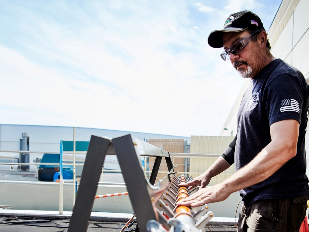 A worker adjusts a set of orange pipes on a support frame, set against a blue sky and industrial background.